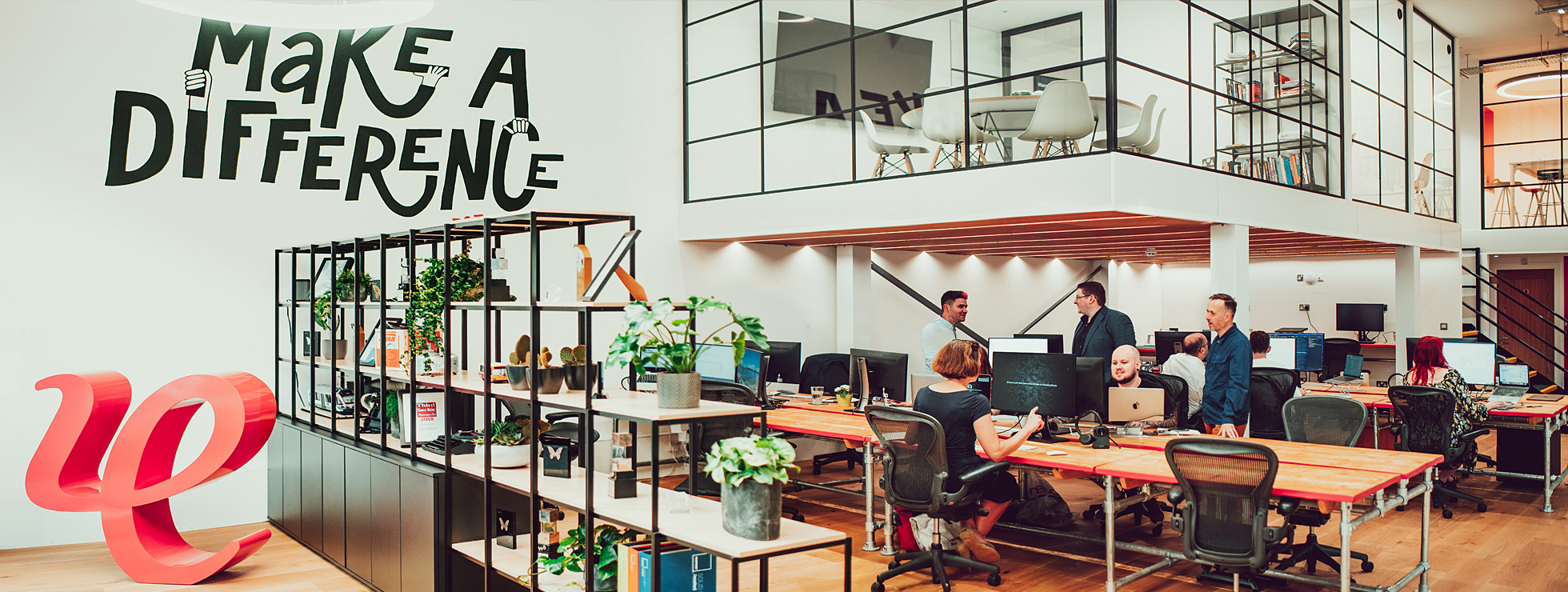A view across the IE Digital studio in Birmingham, showing members of the agency team collaborating on projects. There's a mural on the wall with the agency's motto "Make a difference, est. 1994" and a large red 3D version of IE's logo marque.