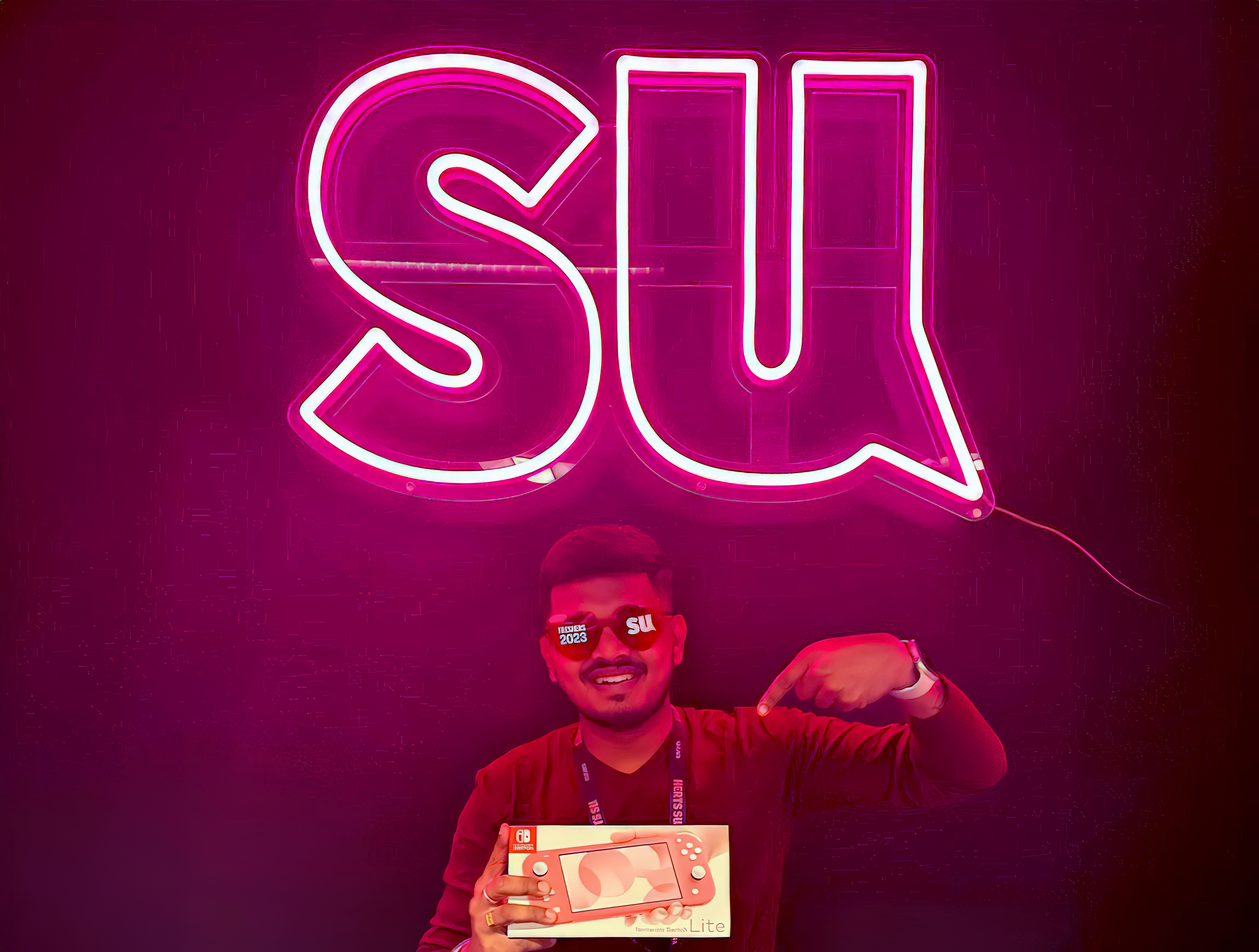 A student holding a boxed Nintendo Switch, part of a giveaway on HertsSU instagram. He's in front of a bright pink neon version of the 'SU' logo, with the speech mark tail on the letter U. He's also wearing branded Herts SU sunglasses and lanyard