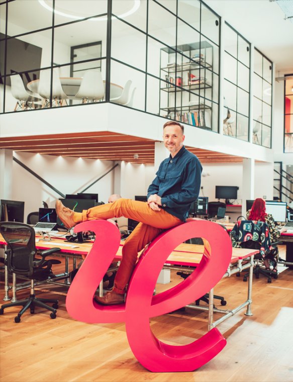 The agency's founder Ollie Leggett (Managing Director and Brand Consultant) sitting atop a large 3D version of the IE logo marque. He's in IE's stunning Birmingham studio.