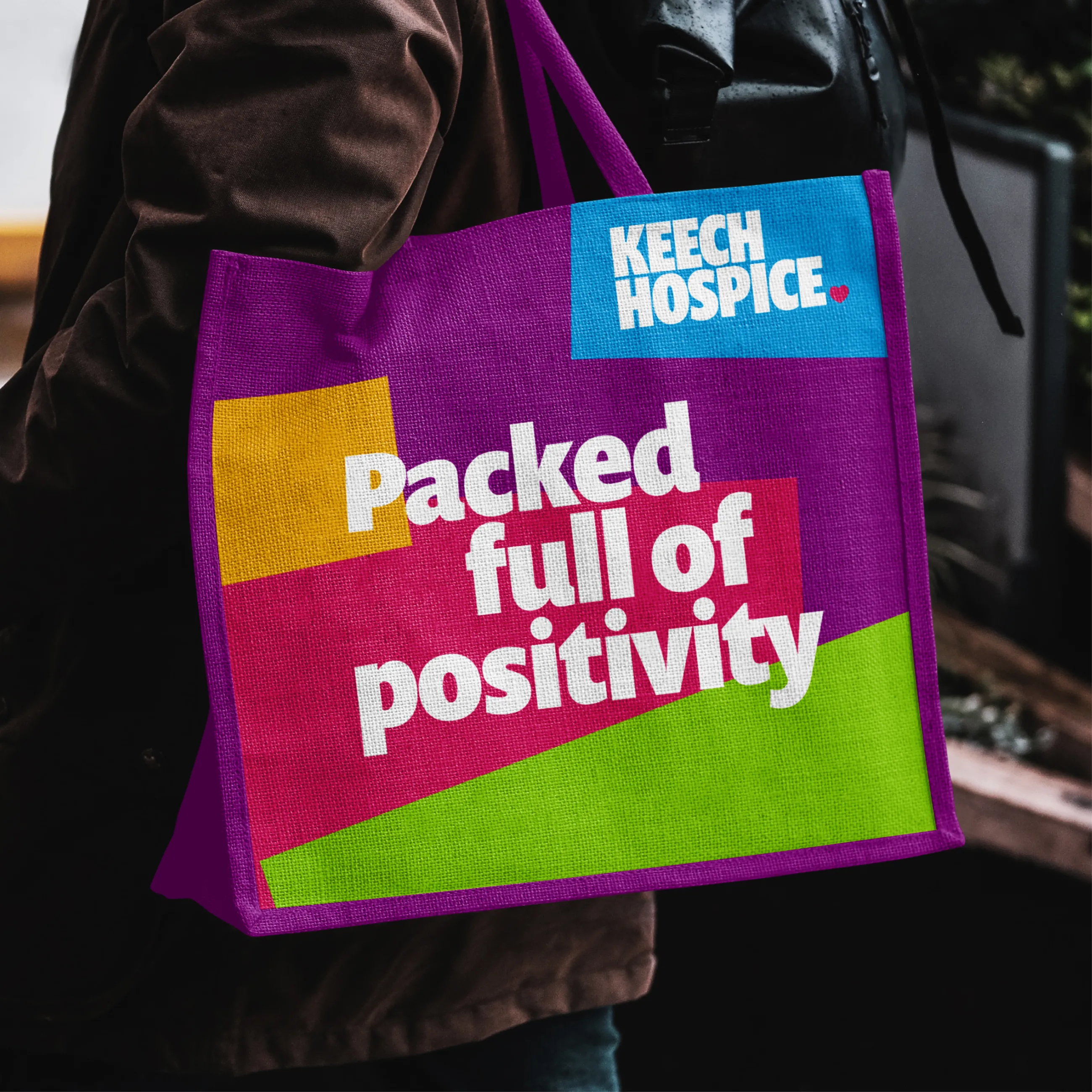A purple tote bag with the Keech Hospice logo with the text saying 'Packed full of positivity' in front of a variety of collaged squares